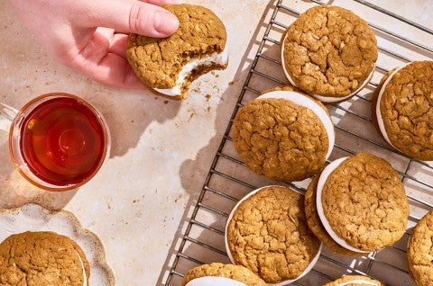 A hand holding an oatmeal cream pie that has a bite taken out of it with others on a cooling rack. - select to zoom