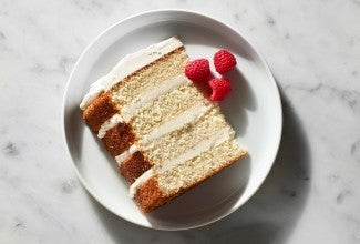A slice of a four-layer Tender White Cake filled with vanilla buttercream and garnished with raspberries on a plate
