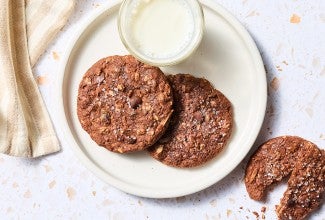 Two Chocolate Lactation Cookies on a plate with a glass of milk and another cookie on the counter next to the plate.