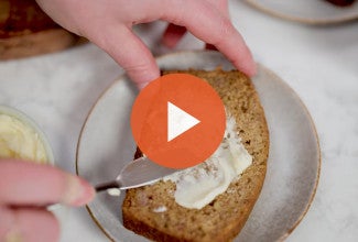 Spreading butter on banana bread with a play button to watch the video. - select to zoom