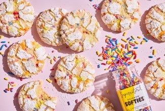 Sprinkle Crinkle Cookies on a light lavender background and a bottle of colorful sprinkle