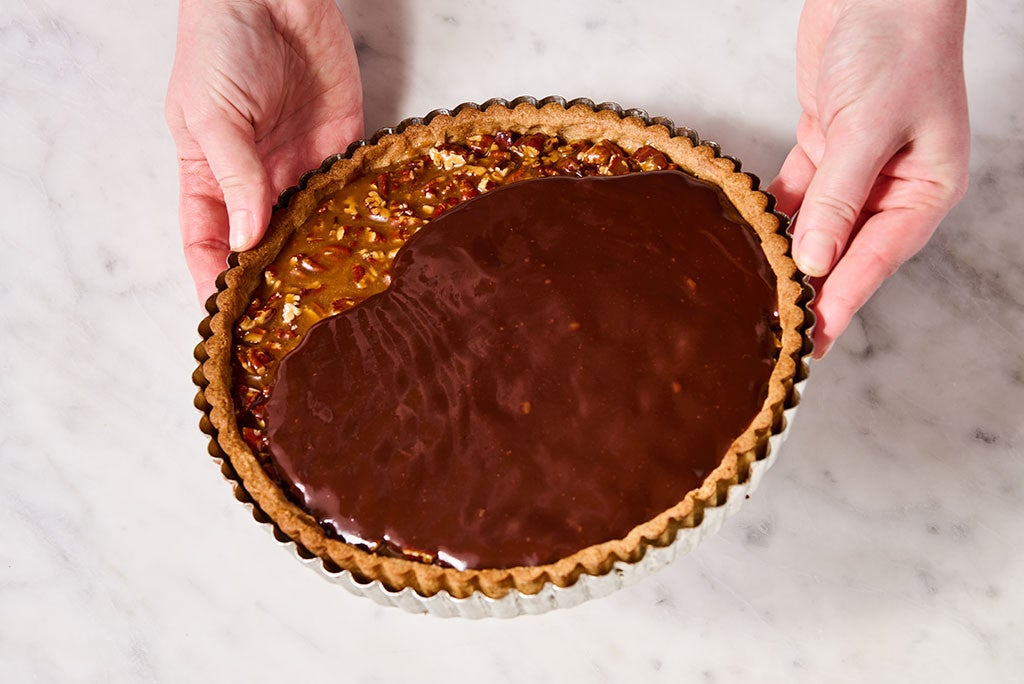A person tilting a round tart pan filled with caramel and chocolate to show spreading out the chocolate glaze evenly