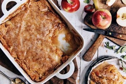 A square ceramic pan of Sugar-Crusted Apple Cobbler with some portioned into a small bowl and a few fresh apples to the side