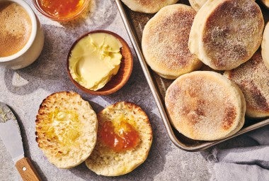 A sliced Sourdough English Muffin on a counter with butter and jam and several others on a sheet tray - seen from above